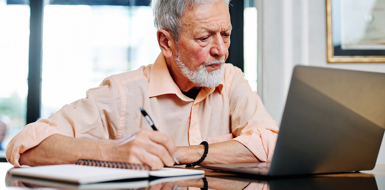 White man with beard looking at computer while writing on a pad of paper with a pen