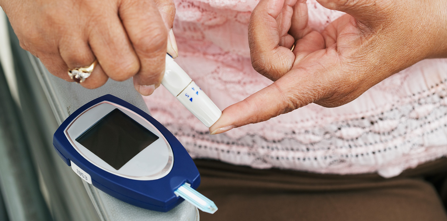 Two hands of an older white lady taking blood sample from her index finger, blood sugar monitor on the table below the hands