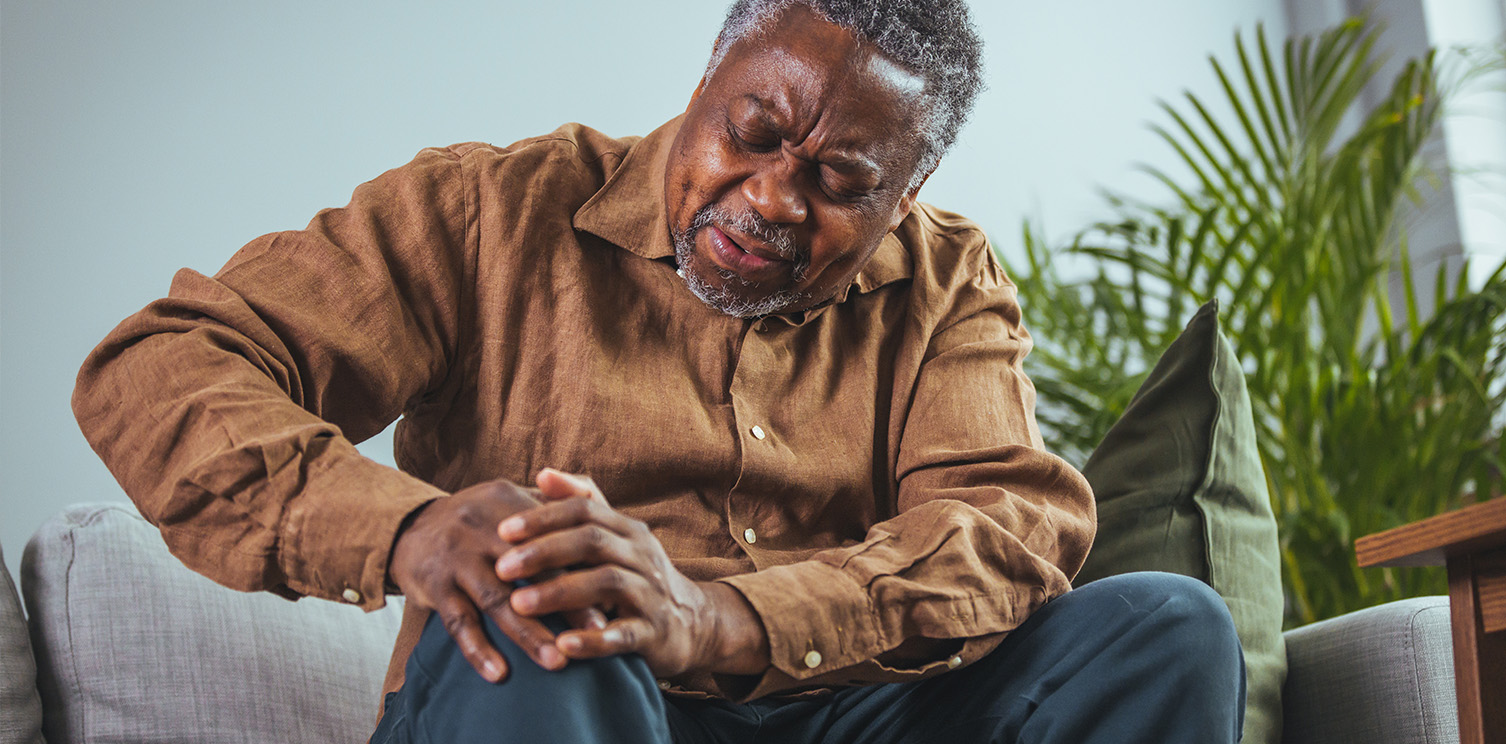 Older black man in rust-colored shirt looking down and holding his knee