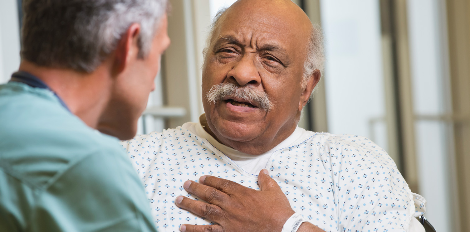 Older black man in hospital gown with his hand on his chest, facing a male doctor