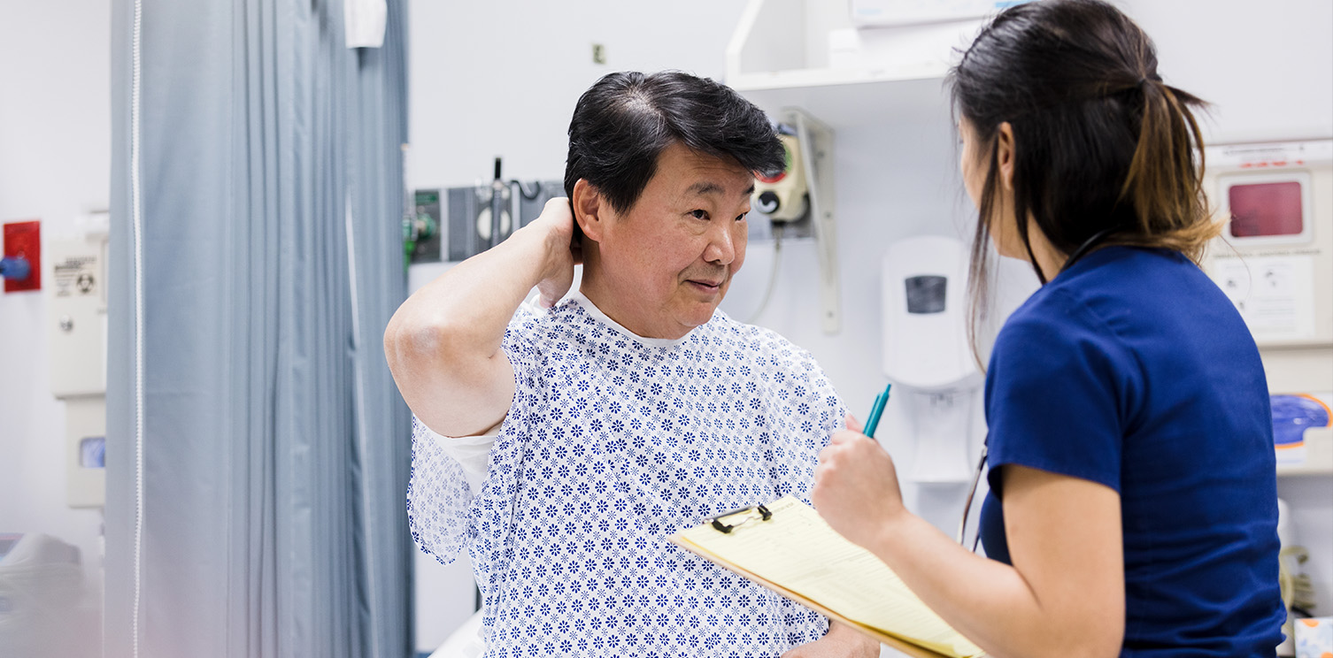 Older Asian man in hospital gown touches back of neck while speaking to female doctor